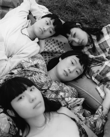 Black and white portrait of four young women lying on a patterned blanket in the grass, gazing directly at the camera with neutral expressions.