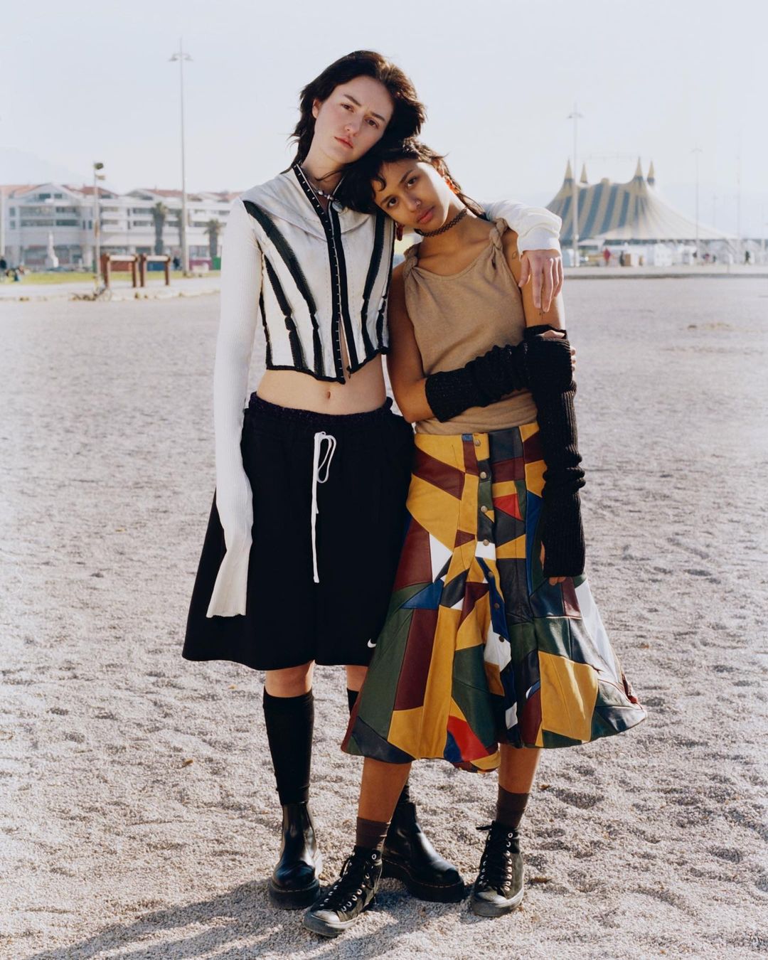 Two young women pose outdoors in fashionable, vintage-inspired clothing. One rests her head on the other's shoulder. A circus tent is visible.