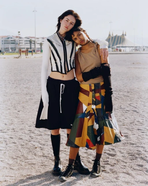 Two young women pose outdoors in fashionable, vintage-inspired clothing. One rests her head on the other's shoulder. A circus tent is visible.