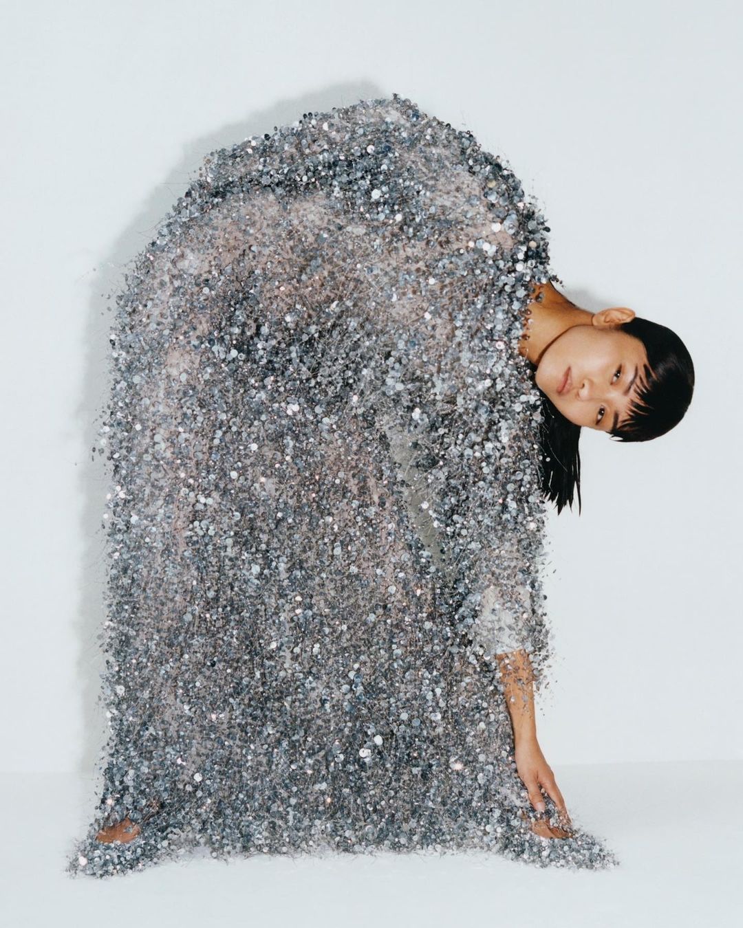 An Asian woman stands draped in a sparkling silver, sequined dress against a white background in a studio portrait, leaning to her right.