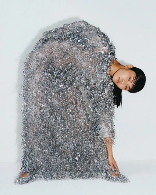 An Asian woman stands draped in a sparkling silver, sequined dress against a white background in a studio portrait, leaning to her right.