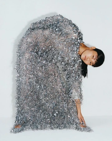 An Asian woman stands draped in a sparkling silver, sequined dress against a white background in a studio portrait, leaning to her right.