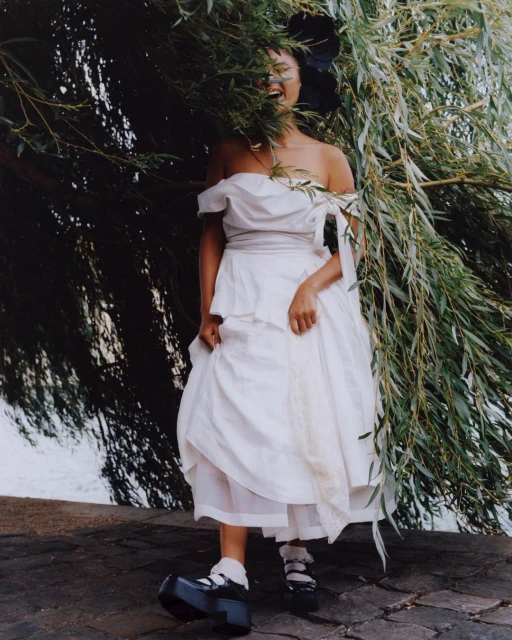 Woman in white dress with black Mary Jane shoes poses next to green weeping willow tree; outdoor fashion editorial photoshoot.