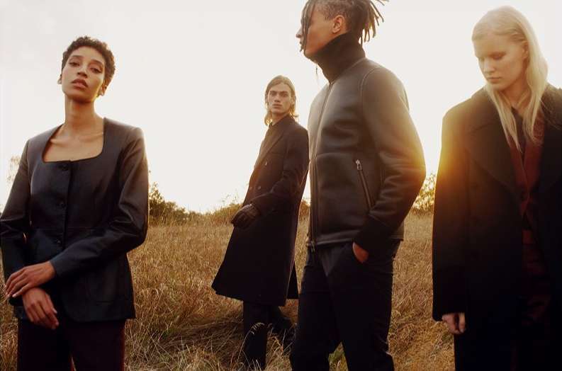 Fashion editorial shot of four models in a field wearing dark coats, exuding a sense of mystery and elegance against a golden sky.