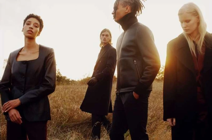 Fashion editorial shot of four models in a field wearing dark coats, exuding a sense of mystery and elegance against a golden sky.