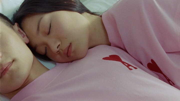 Close-up of a young woman resting her head on a man's chest, both wearing light pink sweatshirts with red heart logos.