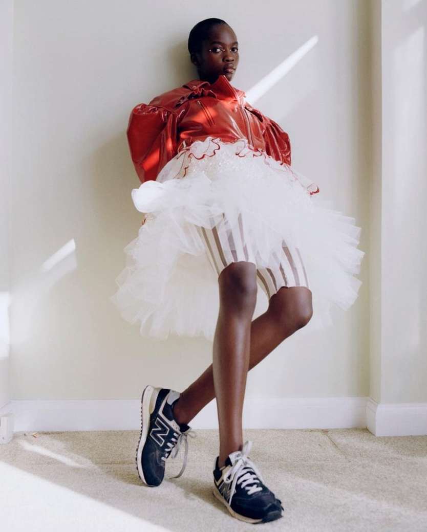 Young Black woman posing in a red jacket and striped skirt with a white tulle overlay, standing against a white wall with shadows.