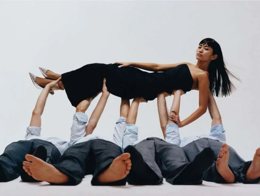 Fashion shot of a woman in a black dress elevated and supported by five prone people in dress shirts and pants on a white background.