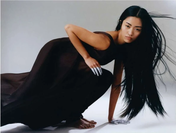 Asian woman with long black hair wearing a dark sleeveless dress and silver fingernails crouched on a white seamless background