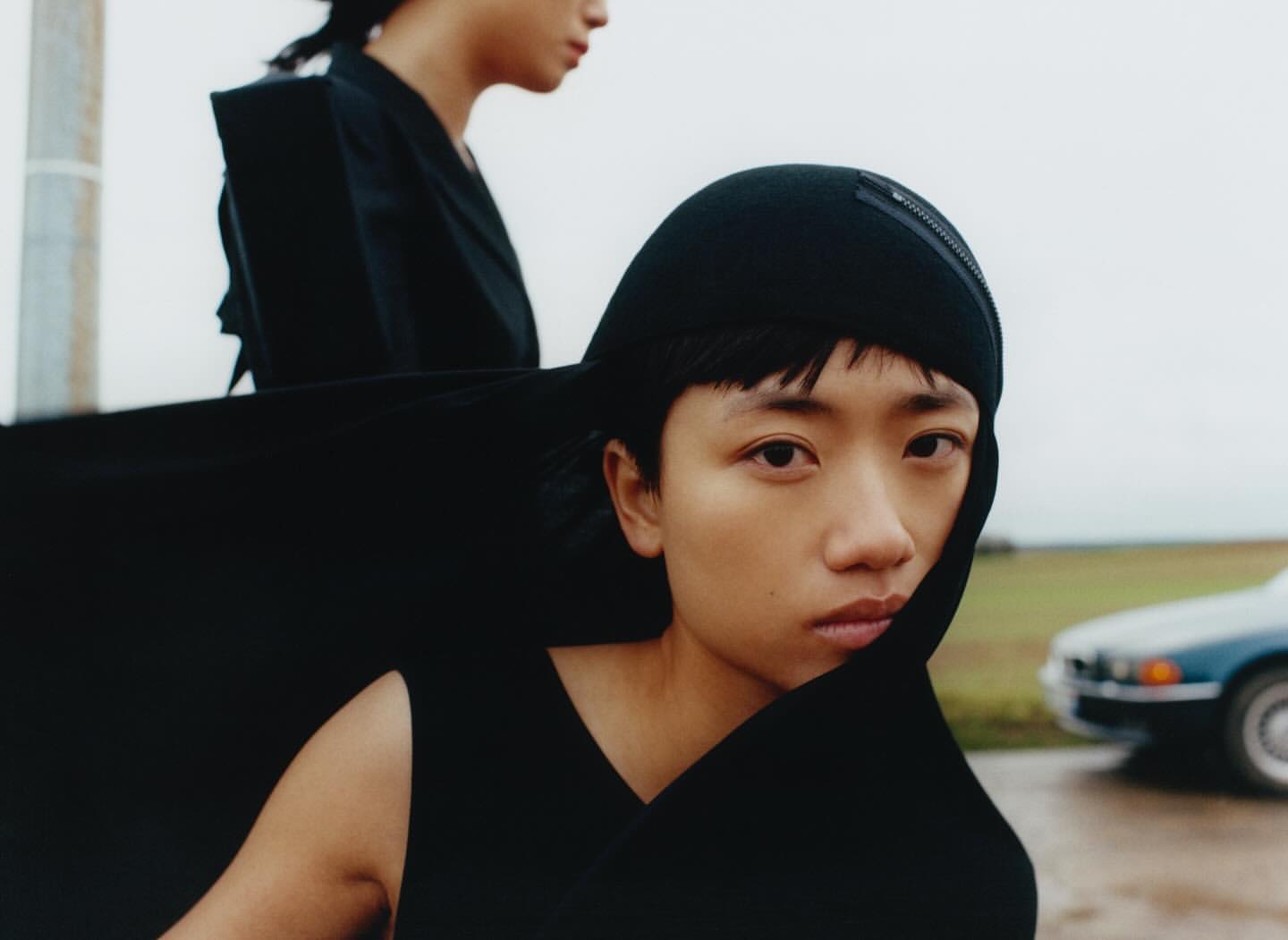 Two Asian women model black avant-garde fashion outdoors in a field with overcast skies and a glimpse of a car; medium shot.