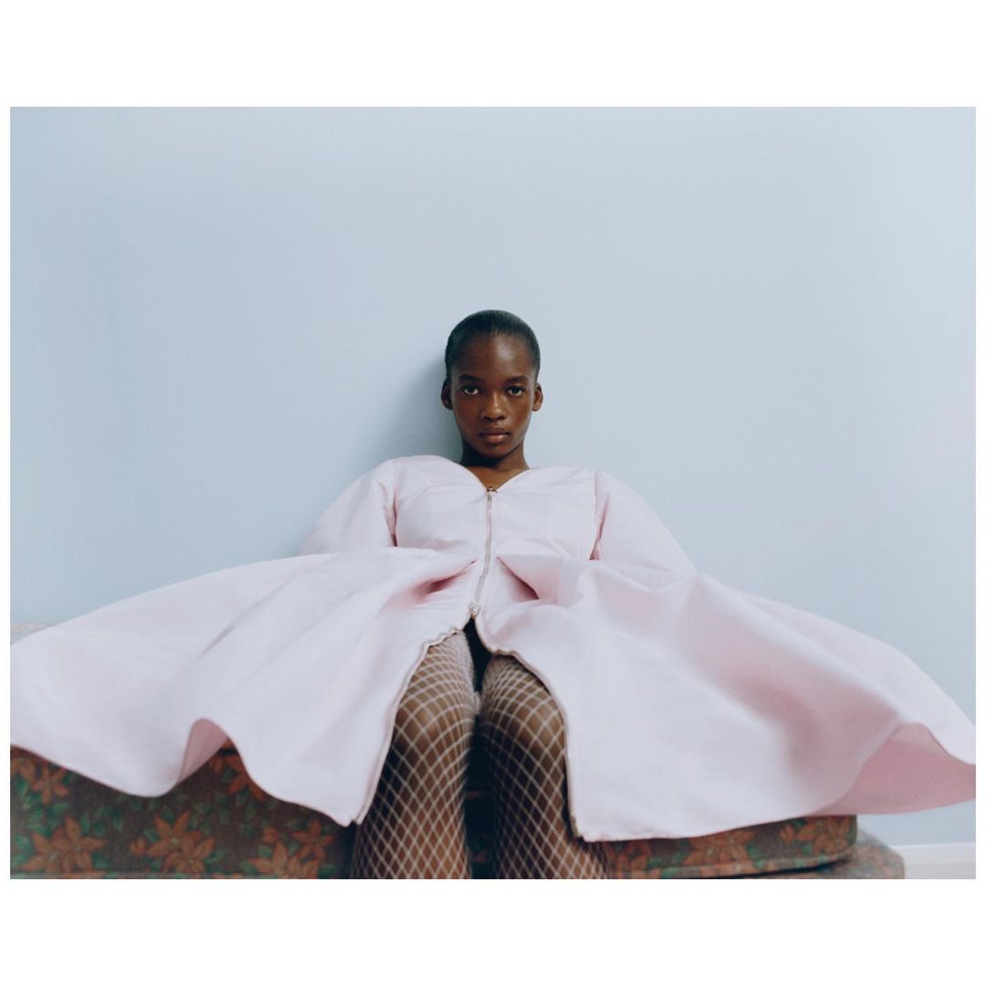 Woman in a pink dress with puffy sleeves and fishnet stockings sitting on a patterned couch against a light blue wall.