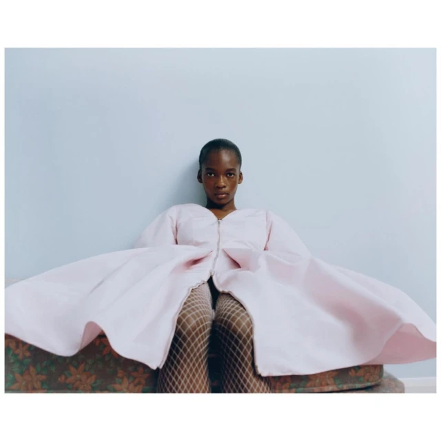 Woman in a pink dress with puffy sleeves and fishnet stockings sitting on a patterned couch against a light blue wall.