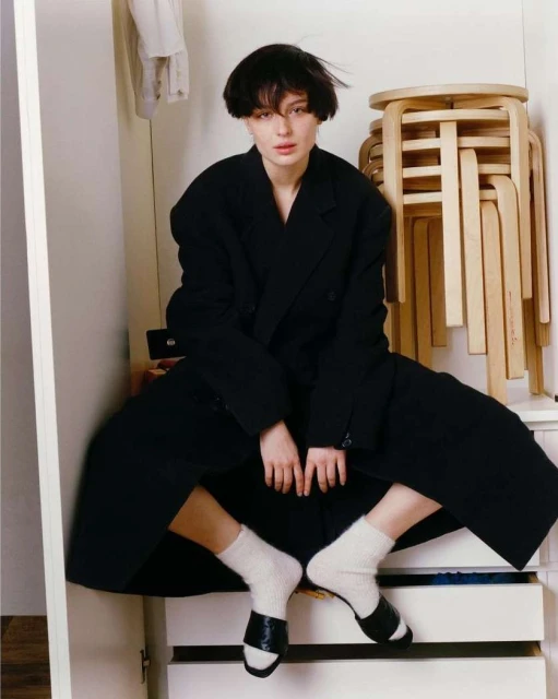 A woman with short dark hair sits in a closet wearing a black coat and white socks with black sandals, surrounded by wooden stools.