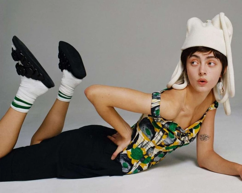 A stylish young adult woman poses on the floor wearing a patterned top, black pants, socks with green stripes, and an unusual white hat.