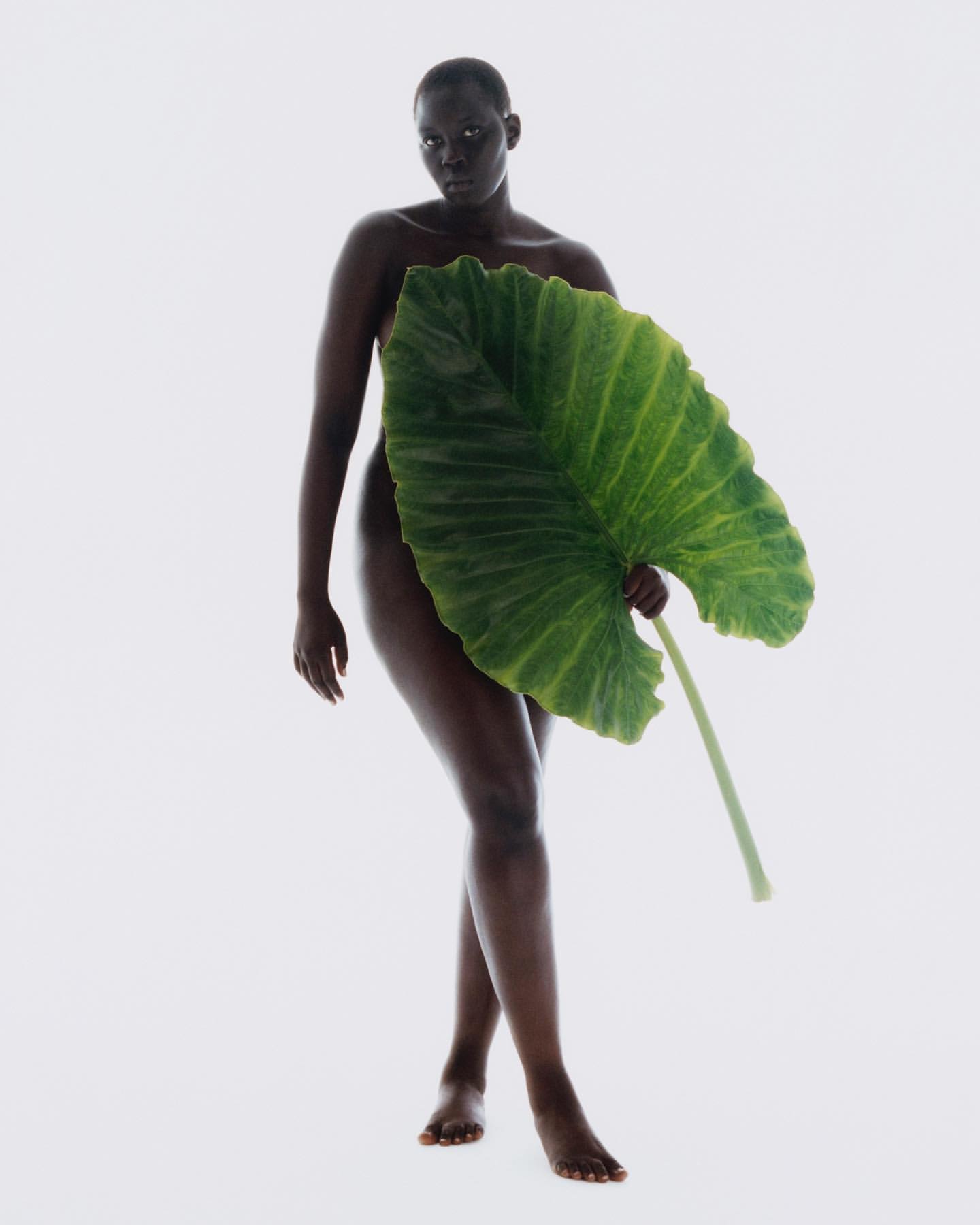 Striking portrait of an adult woman standing in a bright photo studio, partially covered by a large green leaf, creating a natural and artistic aesthetic.