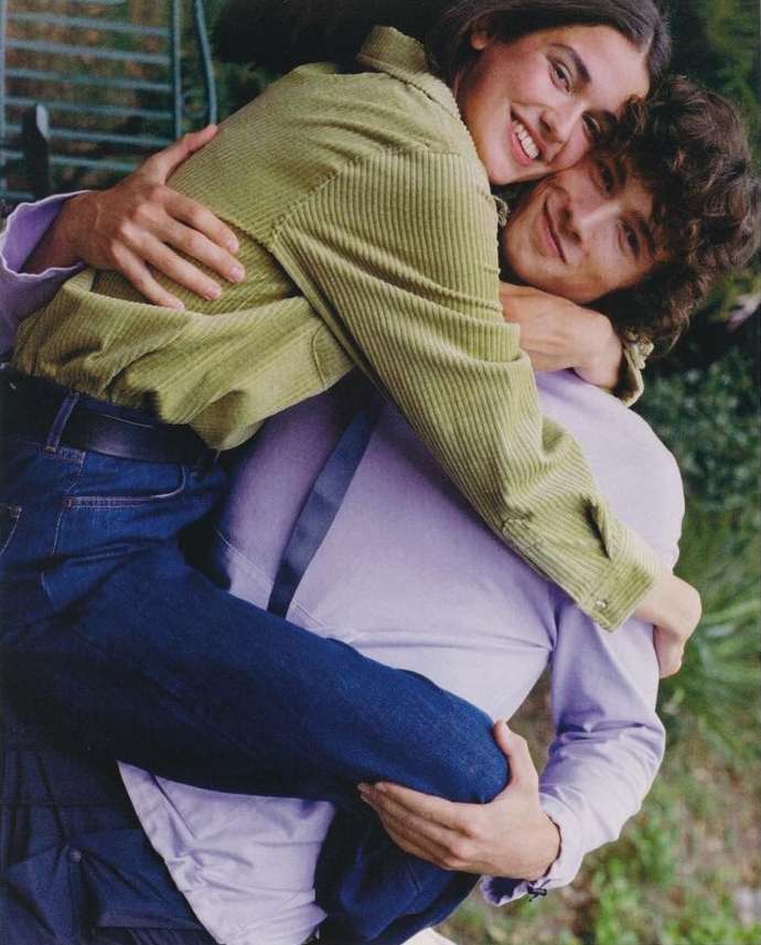 A smiling woman hugs a man outdoors. The woman wears a green corduroy jacket, the man a lilac shirt. They stand amid greenery.