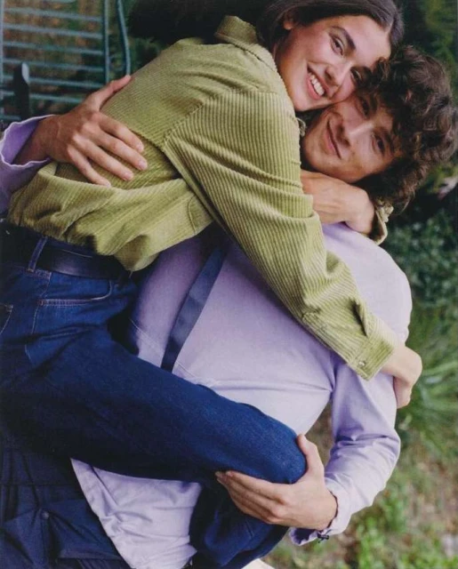 A smiling woman hugs a man outdoors. The woman wears a green corduroy jacket, the man a lilac shirt. They stand amid greenery.