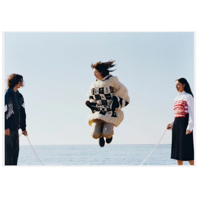 Three people on a beach play jump rope. One person jumps while the other two hold the rope, wearing jackets and casual outfits.