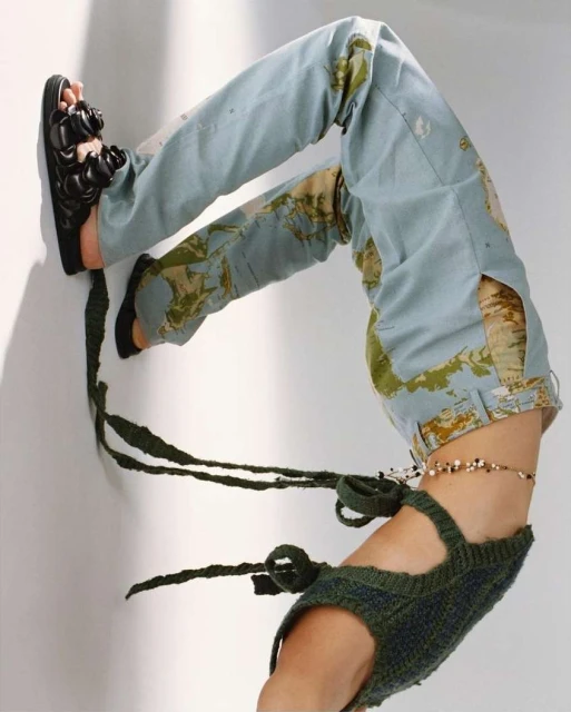 Contortionist wearing map-print pants, black sandals, and green knit top, bending backwards against a white wall in a studio setting.