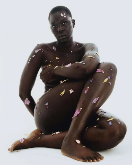 Striking nude portrait of a dark-skinned adult female model adorned with delicate flower petals against a clean white background.