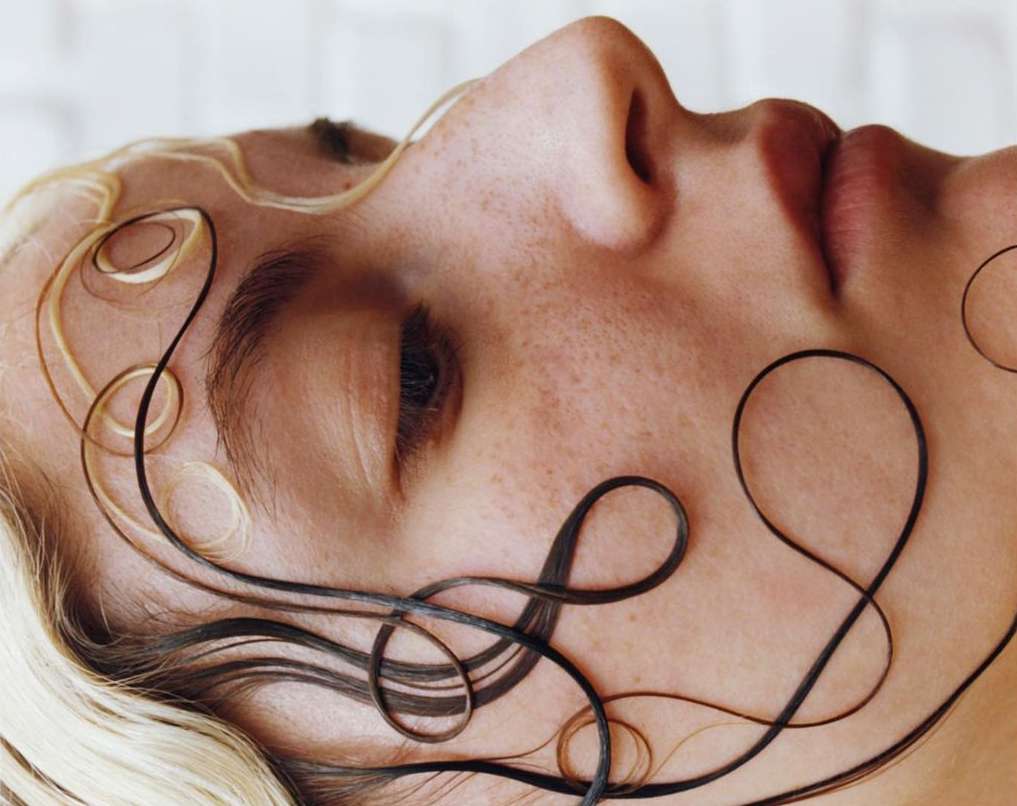 Close-up portrait of a woman with blonde hair, lying down. Dark-brown hair strands are styled in curved lines across her face.