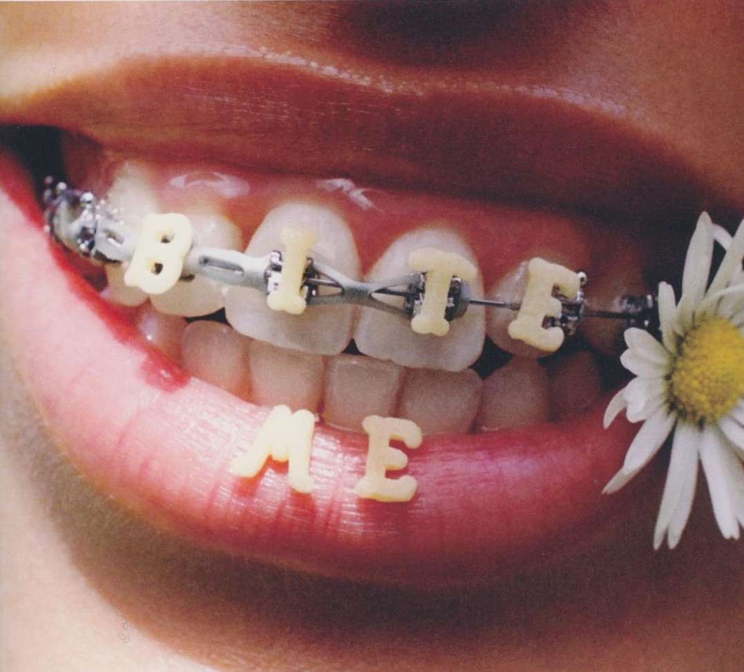 Close-up of lips and teeth with braces spelling 'Bite Me' in letter-shaped pasta, adorned with a daisy, on a neutral background.