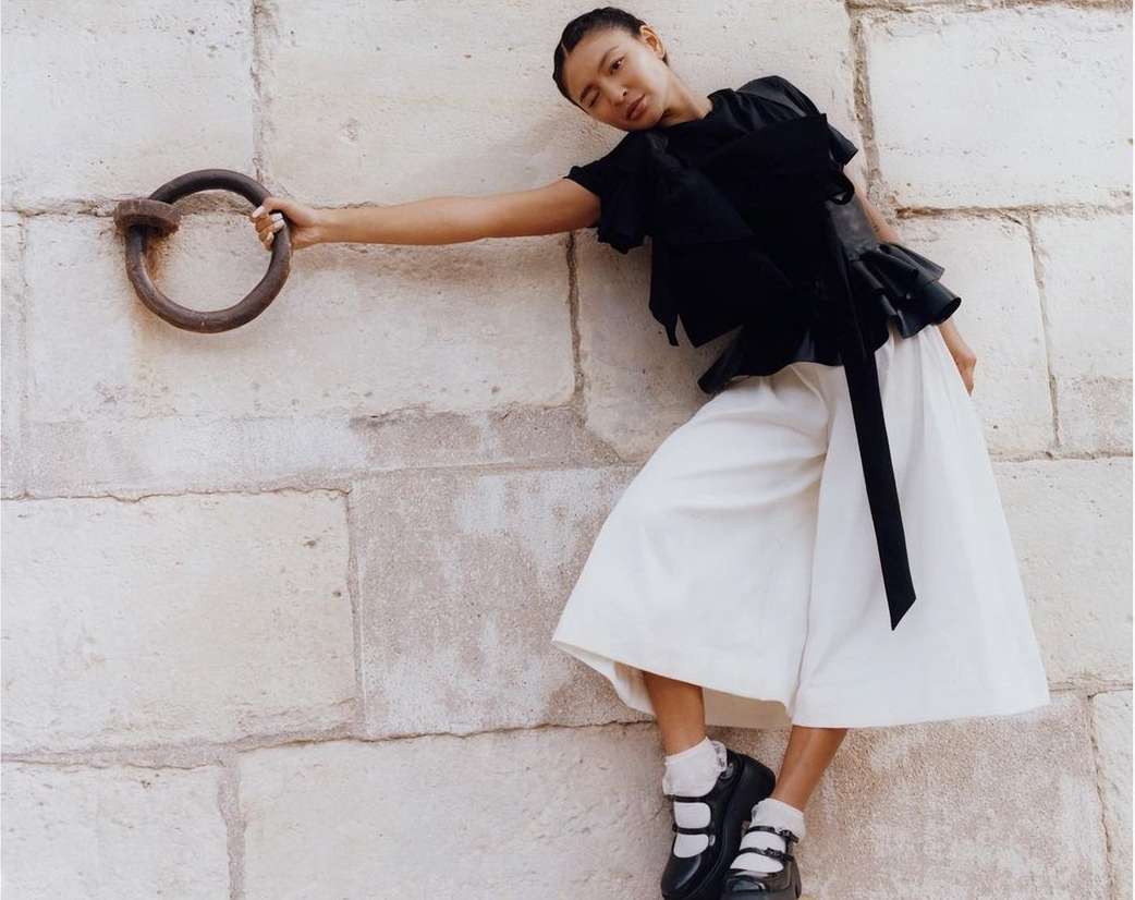 Fashion shot of an Asian woman in a black top and white culottes, leaning against a stone wall and holding a metal ring.