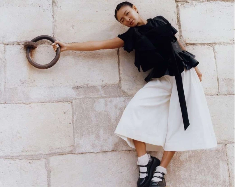 Fashion shot of an Asian woman in a black top and white culottes, leaning against a stone wall and holding a metal ring.