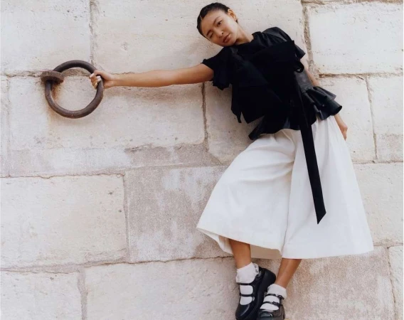 Fashion shot of an Asian woman in a black top and white culottes, leaning against a stone wall and holding a metal ring.