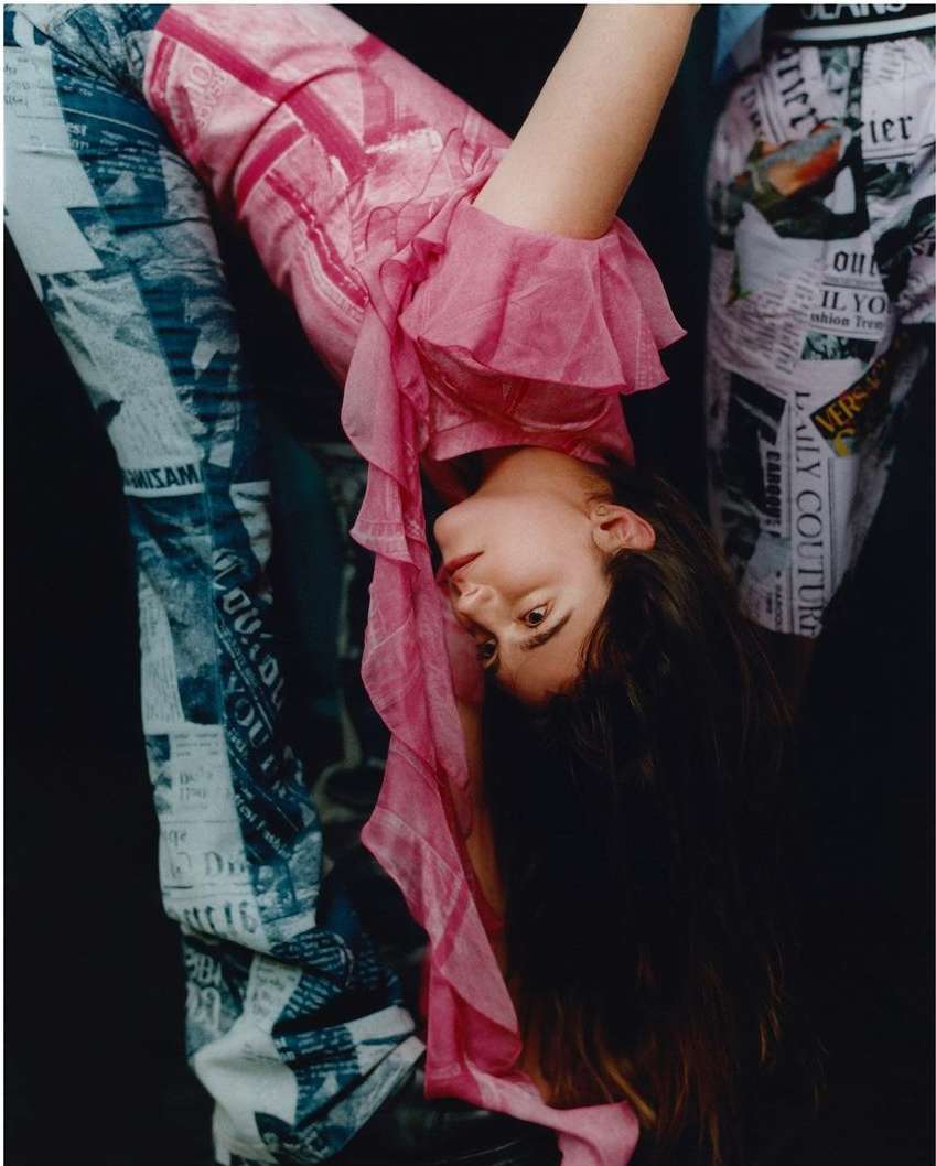 A woman in a pink top and newspaper-print pants bends backwards, viewed from a low angle against a dark, shadowed background in a fashion shot.