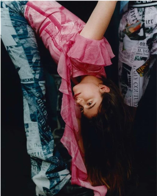 A woman in a pink top and newspaper-print pants bends backwards, viewed from a low angle against a dark, shadowed background in a fashion shot.