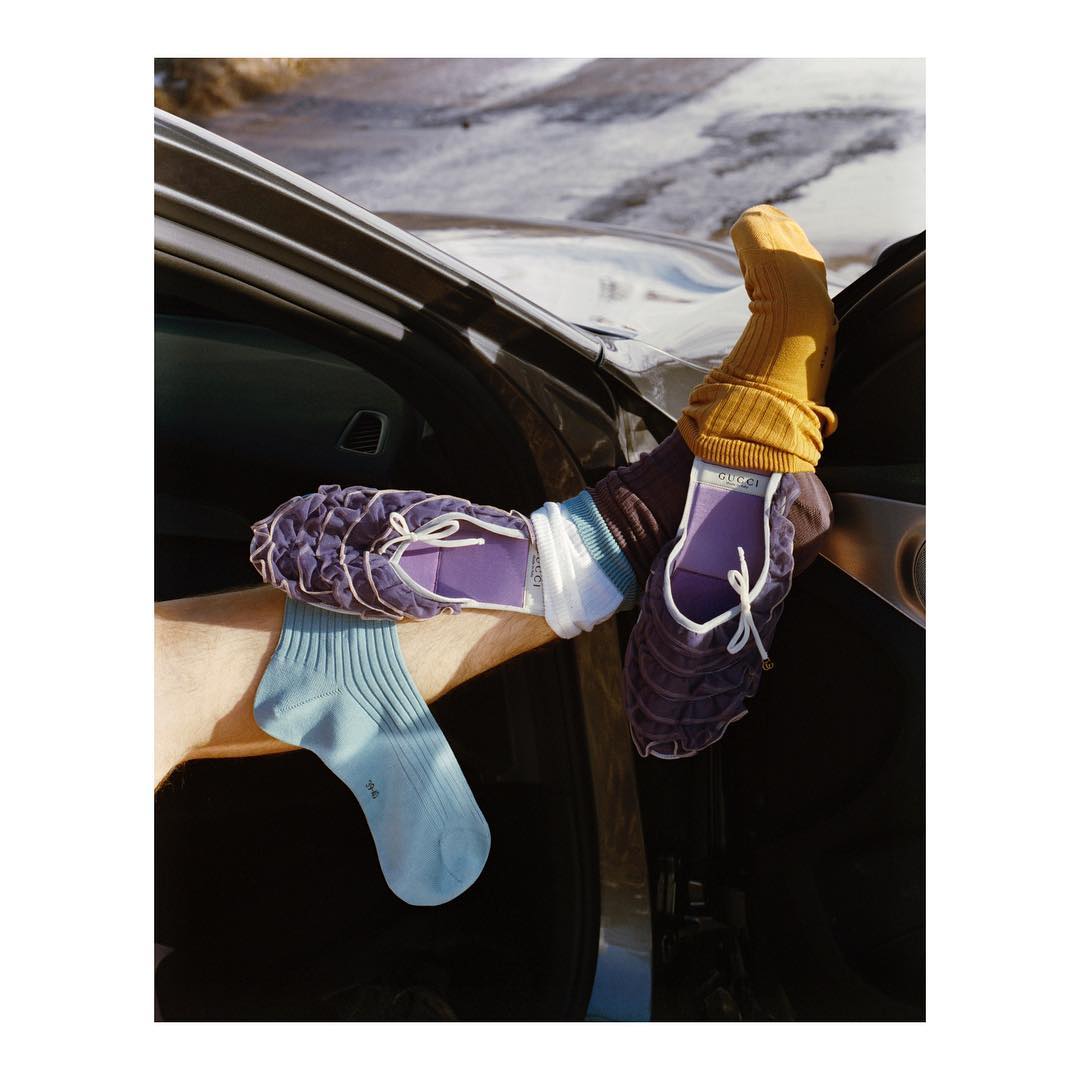 Fashion shot showcasing two arms extending from a black car door, each modeling distinct purple and gold Gucci slipper and sock combinations.