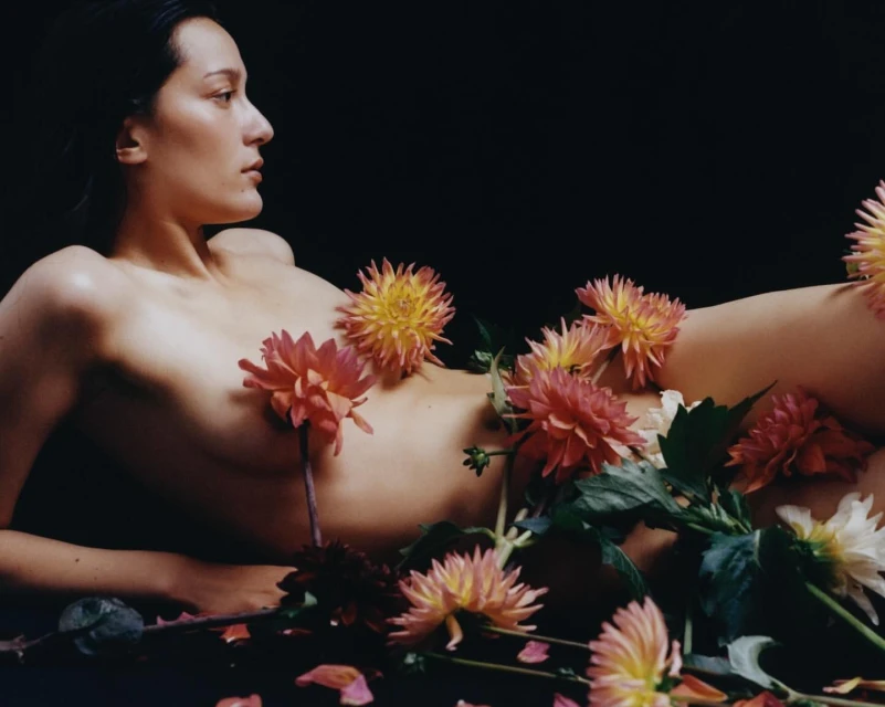 Nude woman lies among pink, peach, and white dahlia flowers against a dark background in a fine art photography studio setting.