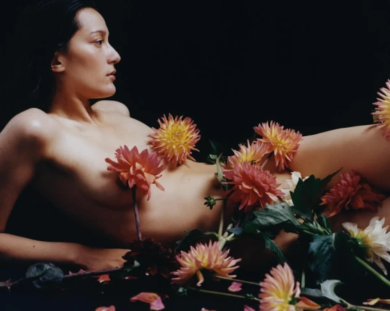 Nude woman lies among pink, peach, and white dahlia flowers against a dark background in a fine art photography studio setting.