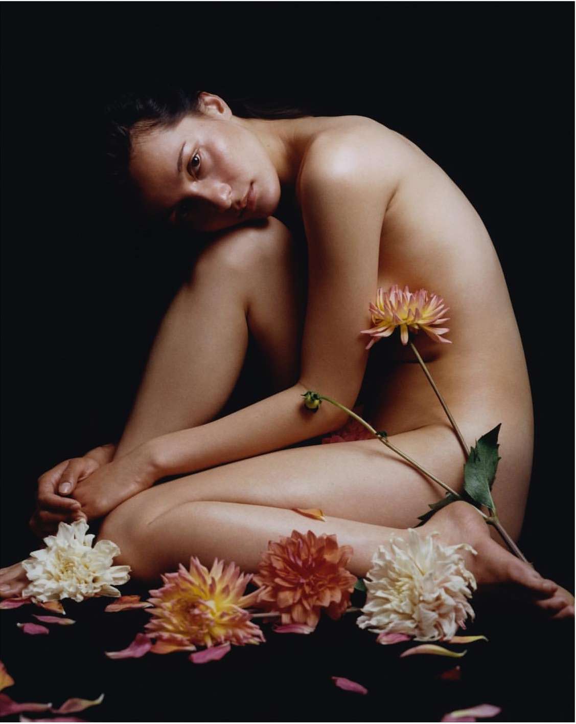 Nude female model poses in studio with flower, surrounded by pink and white petals on dark background in sensual portrait photo.