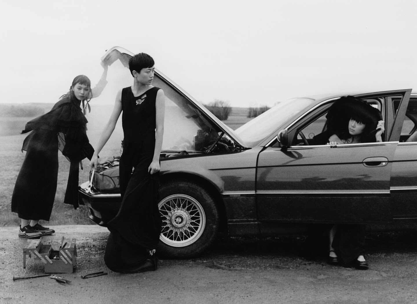 Black and white shot of three women near a vintage sedan with the hood up on a rural road surrounded by grass and trees in overcast lighting.