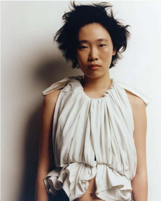 An Asian woman with dark, short hair poses in a draped, light-colored top against a light wall in a close-up portrait shot.