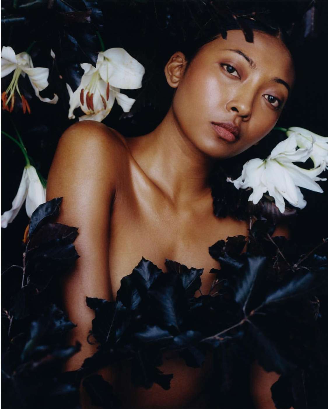 Portrait of a young woman with tanned skin surrounded by white lilies and dark foliage in a studio shot with dramatic lighting.