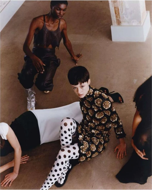 Fashion editorial shot with multiple women wearing designer clothing, seated on a light brown surface. Shot with studio lighting.