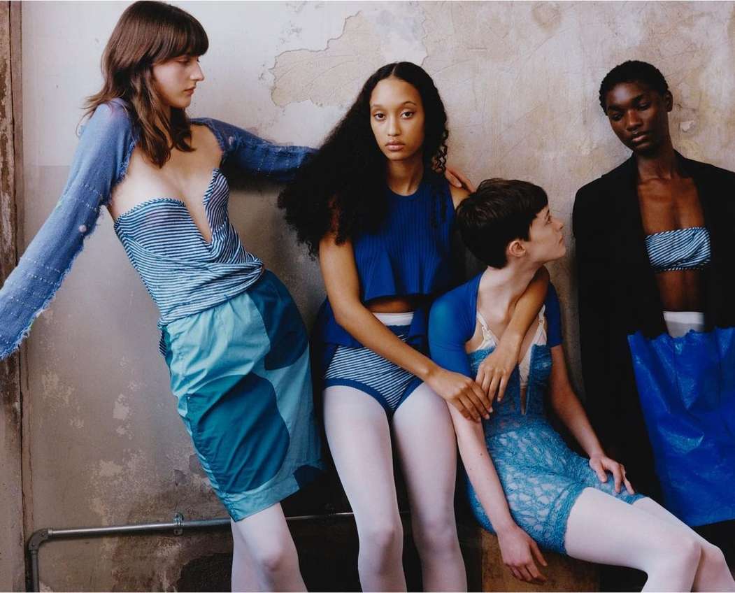 Four diverse women in blue and white fashions pose against a distressed wall in a studio fashion shot showcasing unique designs.