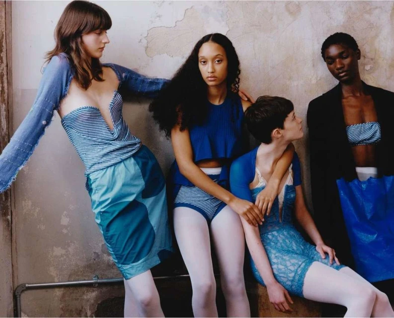 Four diverse women in blue and white fashions pose against a distressed wall in a studio fashion shot showcasing unique designs.