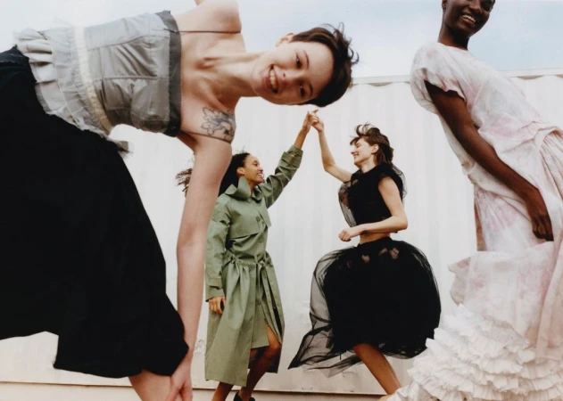 Four women in diverse stylish outfits dancing and posing together outdoors under bright natural daylight.