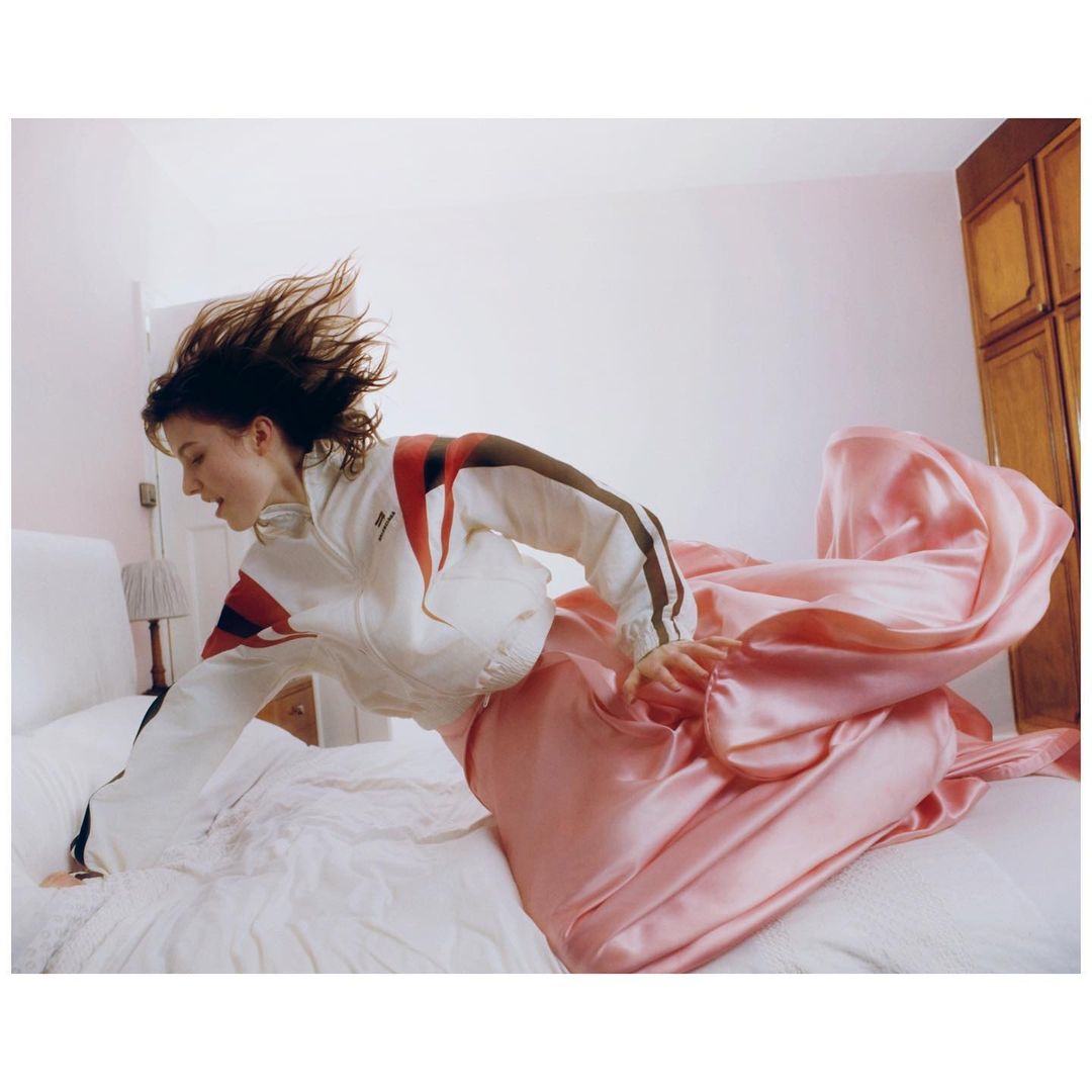 Woman leaps on white bed wearing a white athletic jacket and a pink satin gown, indoor bedroom setting, light and airy aesthetic.