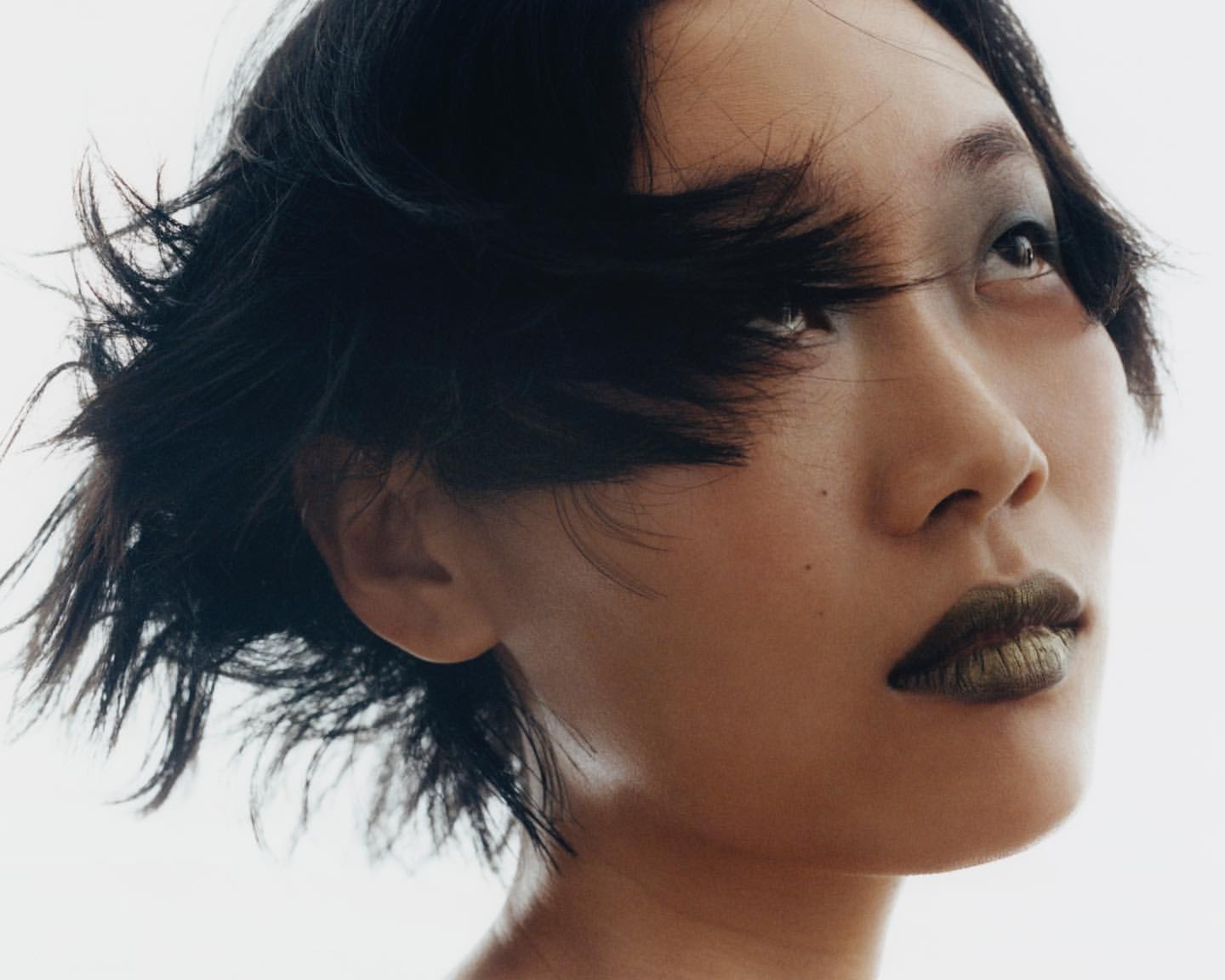 Portrait of Asian woman with short dark hair, wearing green lipstick, looking up against a light background in a beauty headshot.