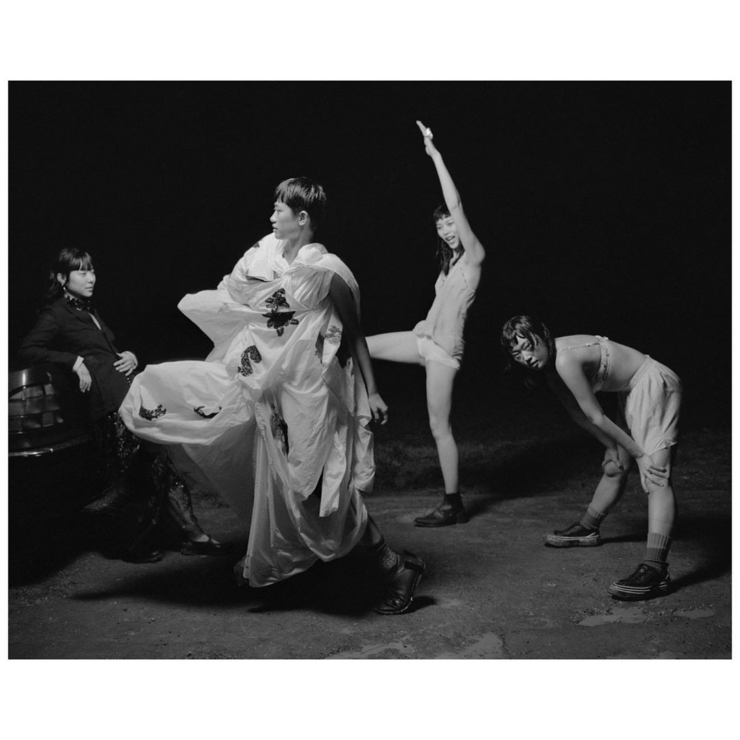 Four young women pose outdoors at night in a grayscale fashion shot; one wears a sculptural white dress and boots, three in casual clothes.