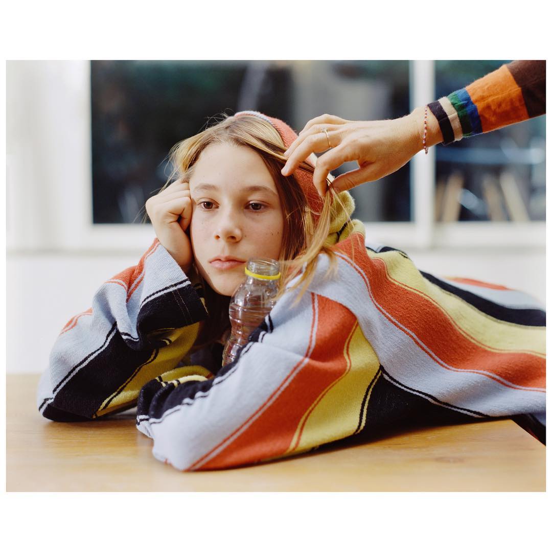 A young person rests at a table wearing a striped hoodie, with an adult's hand gently touching their hair. A water bottle sits nearby.
