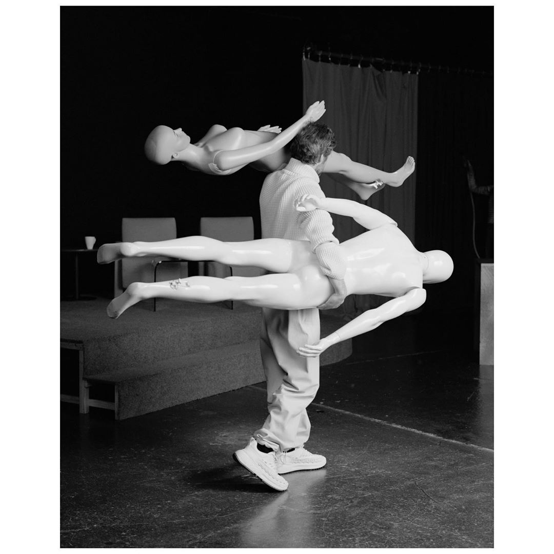 Monochrome shot of a man carrying two white mannequin bodies, one draped across his shoulders and the other supported on his back.