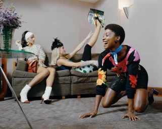 Three models posing in a living room, with a laughing black woman kneeling on the carpet in the foreground.