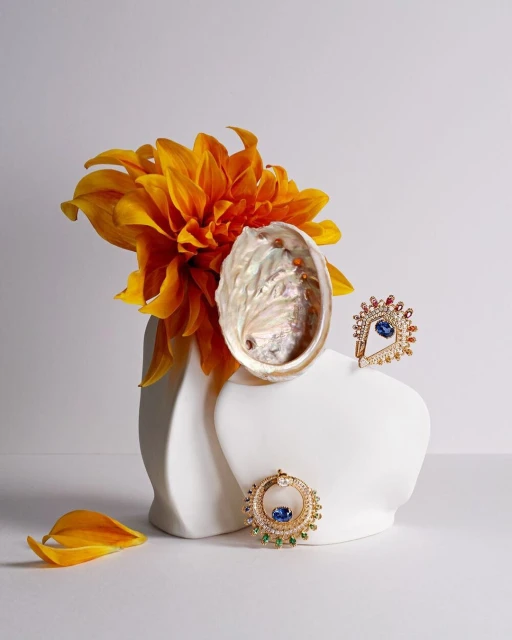Still life featuring an abalone shell, yellow dahlia in white vase, and jewelry with blue gems on white background in studio lighting.