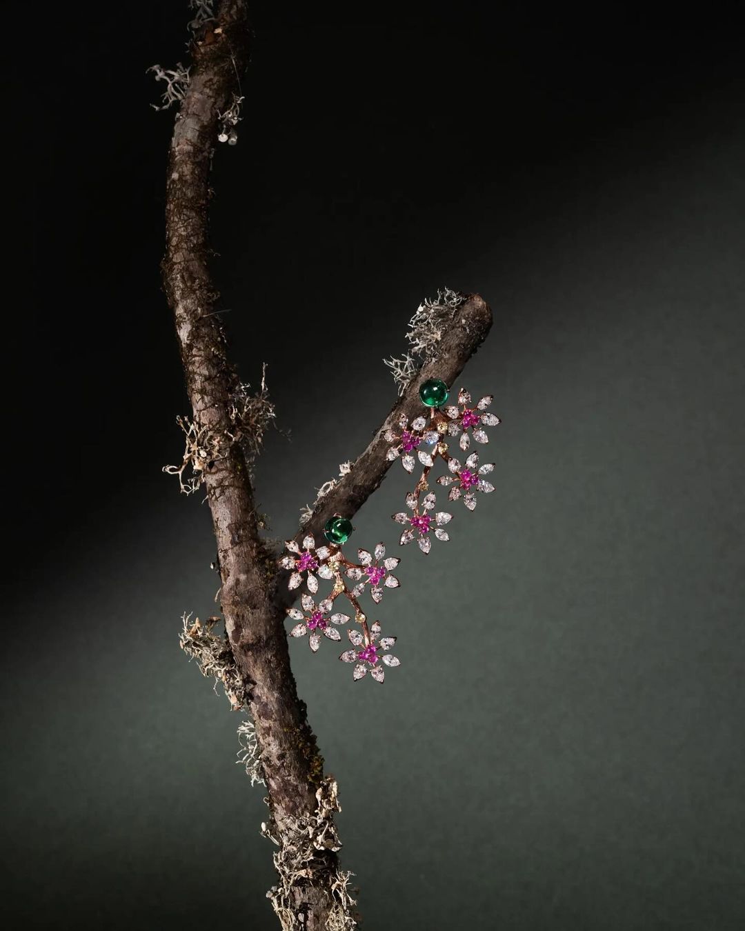 Elegant floral-themed earrings with rubies, diamonds, and emeralds, showcased on a moss-covered branch against a dark background.